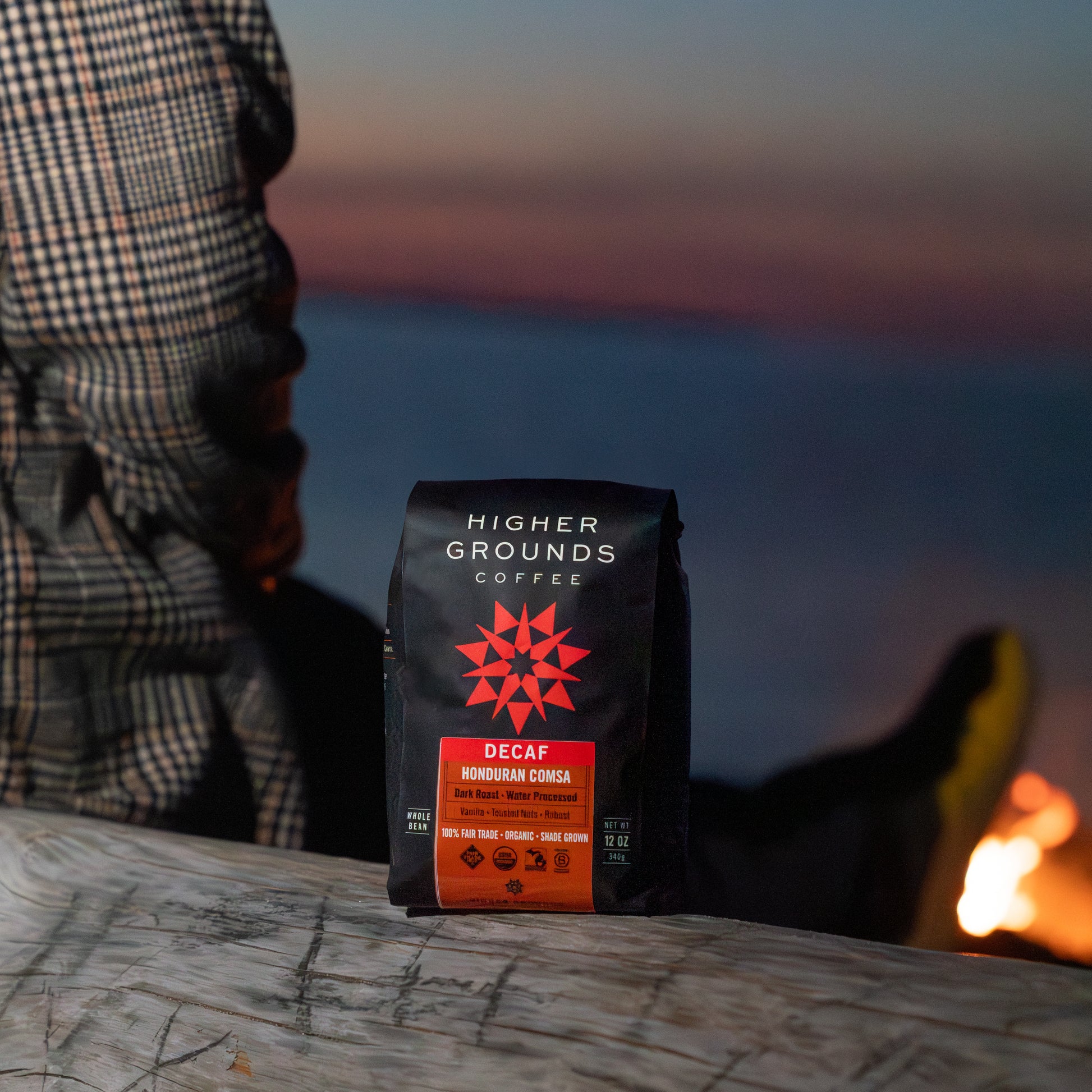 Higher Grounds Coffee Decaf Dark Roast bag on a log with a sunset or sunrise in the background