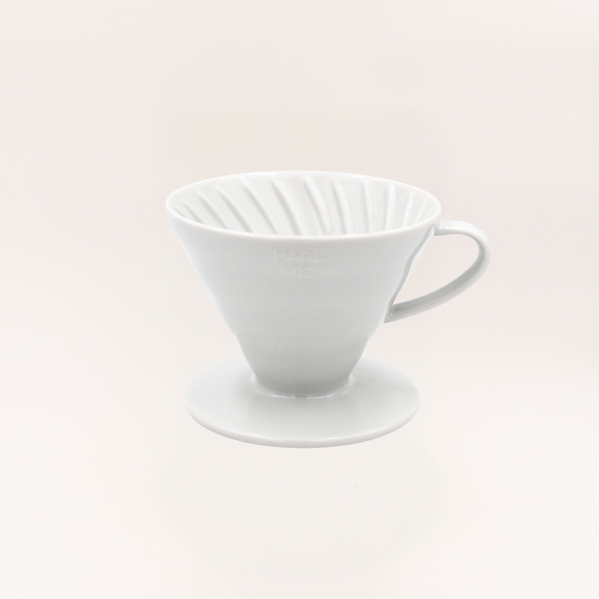 A white ceramic pour-over coffee dripper with a conical shape, ribbed interior, and side handle, resting on a matching saucer against a warm off-white background.